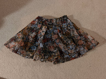 A pleated skirt with a dense floral pattern in muted tones of blue, red, orange, and beige, laid flat on a textured beige carpet.