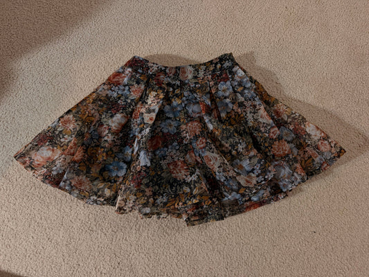 A pleated skirt with a dense floral pattern in muted tones of blue, red, orange, and beige, laid flat on a textured beige carpet.