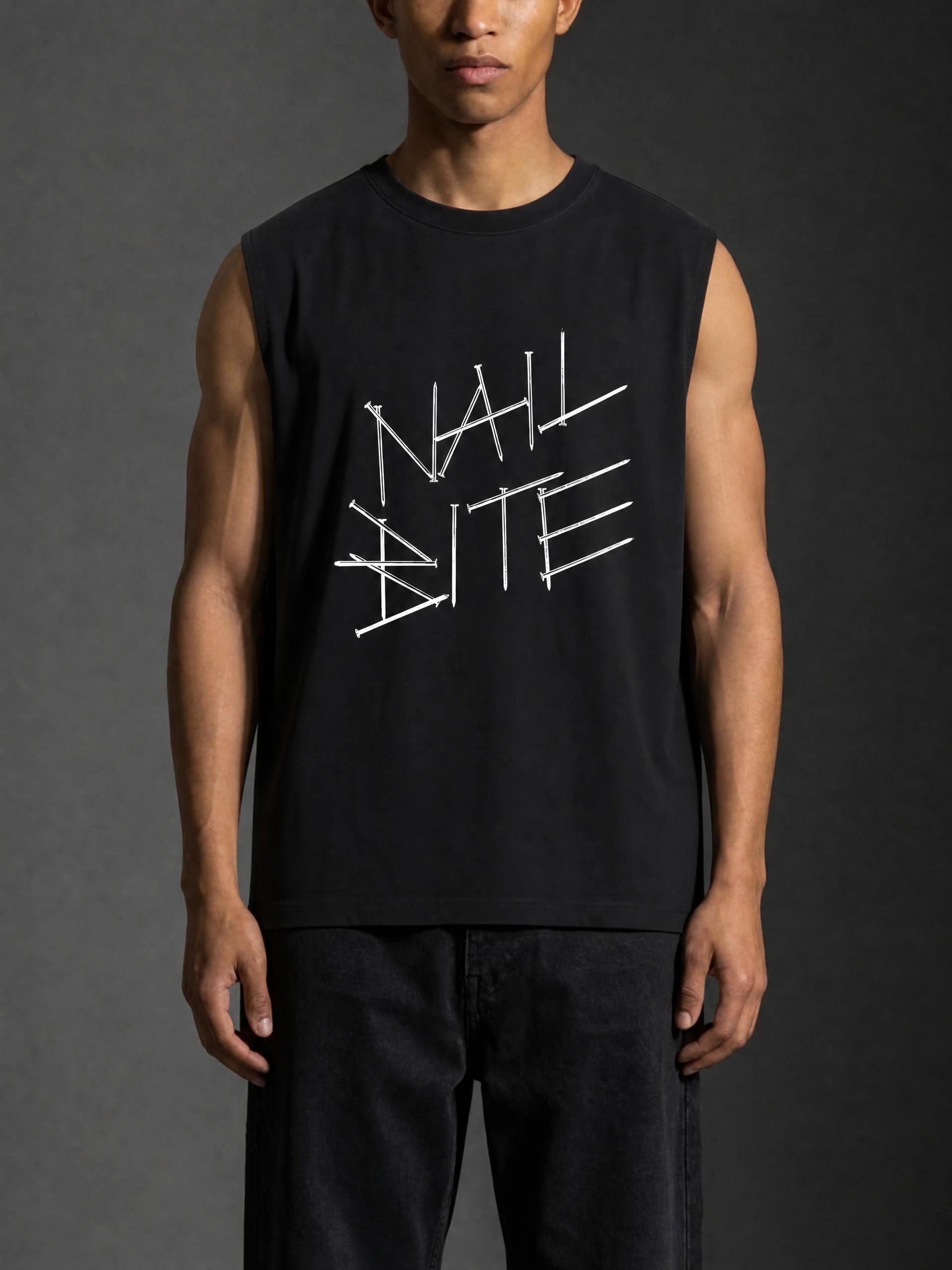 A man wearing a black sleeveless muscle tank top with a white graphic text print reading 'NAIL BITE' across the chest, paired with dark jeans, set against a dark gray studio background.