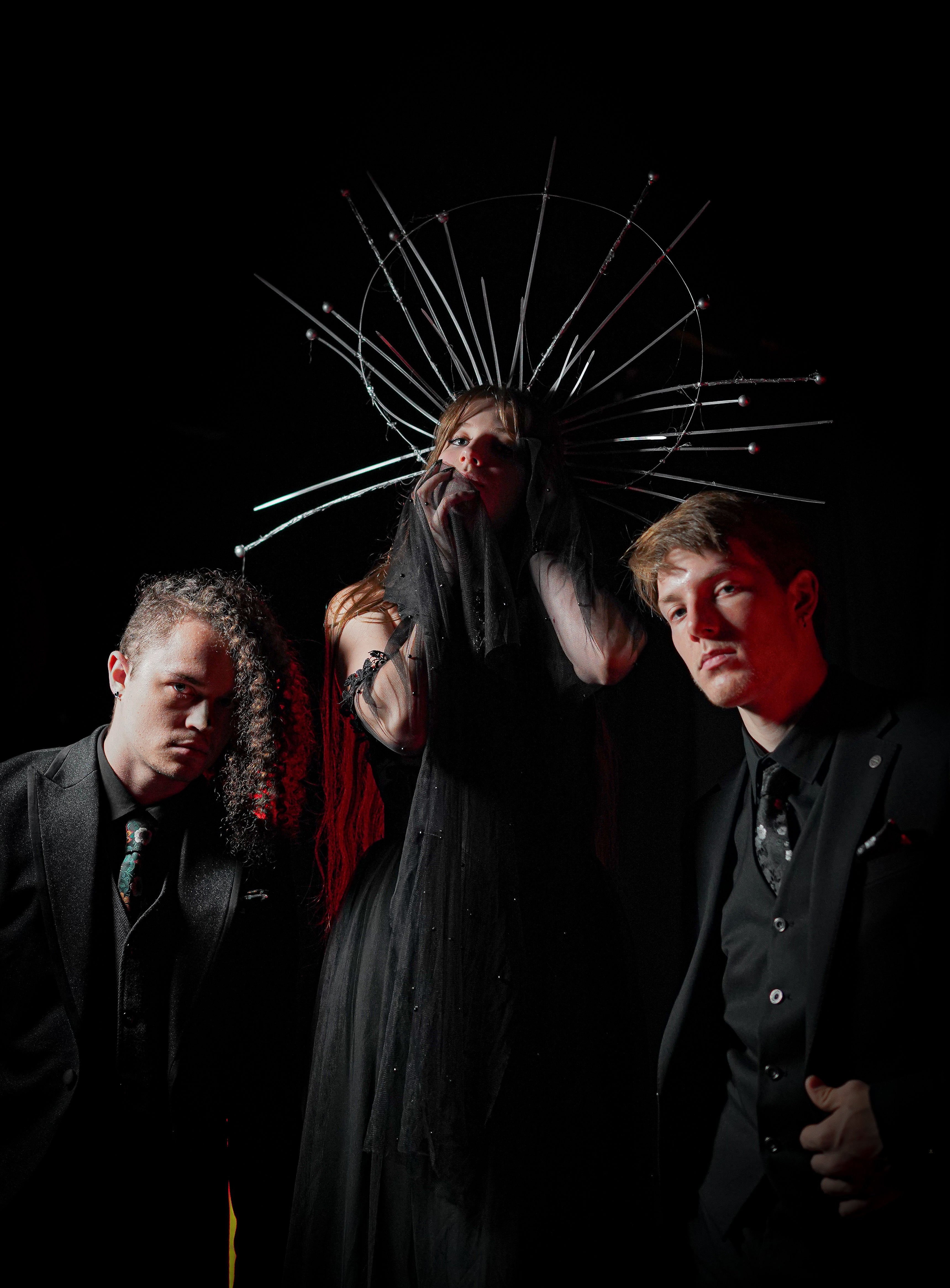 Three men in black clothing posed against a black background. The central figure wears a long black robe and a large metallic halo-like headpiece with many thin rods, while the two men on the sides wear dark suits and ties.